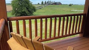 Una terraza de madera con una silla y barandilla, con vista a un campo cubierto de hierba y un edificio distante.