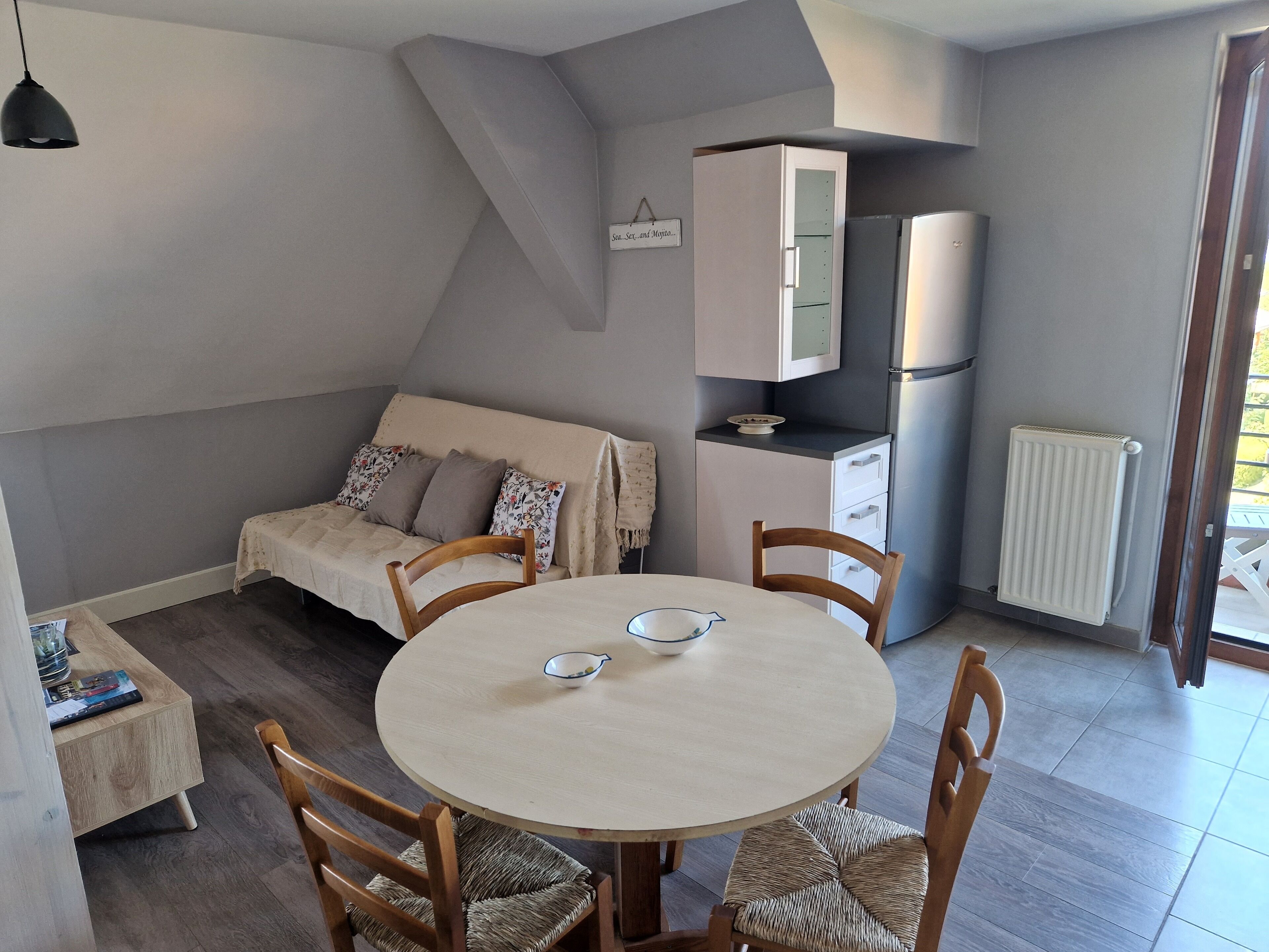 Un espacio habitable compacto con una mesa de comedor redonda, dos sillas de madera, un sofá con cojines estampados, una kitchenette con refrigerador y un radiador.