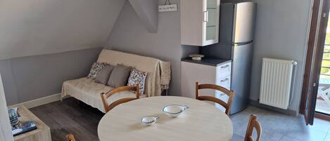 Un espacio habitable compacto con una mesa de comedor redonda, dos sillas de madera, un sofá con cojines estampados, una kitchenette con refrigerador y un radiador.