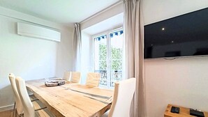 A dining area with a wooden table, white chairs, a TV on the wall, and a balcony with a view. - Viva Riviera 3 bedrooms Natural Loft Luxury Collection (Cannes)
