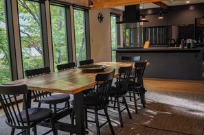 A modern dining area with a wooden table and black chairs, large windows with a view of trees, and a kitchen area with dark cabinetry and appliances.