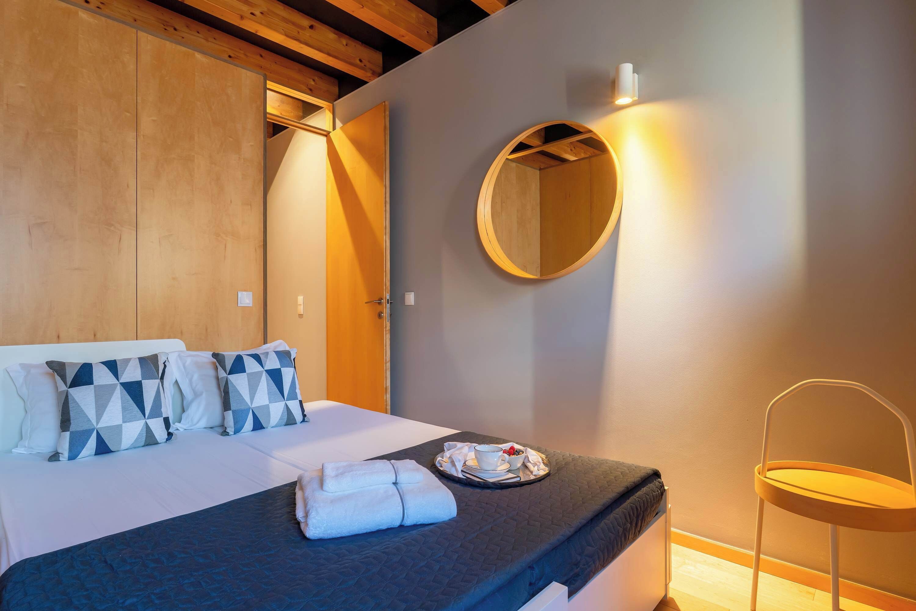 Cama bem arrumada com edredom escuro, almofadas estampadas com padrão geométrico e bandeja com xícara e comida.