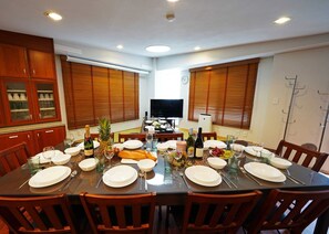 Un coin repas avec une grande table ronde dressée pour six personnes, entourée de chaises en bois. On aperçoit un téléviseur posé sur un meuble au fond de la pièce, et les fenêtres sont équipées de stores en bois.