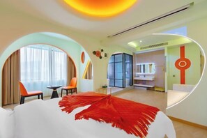 Una habitación de hotel moderna con una cama grande, una mesita pequeña, dos sillas, un escritorio y un baño con un espejo amplio.