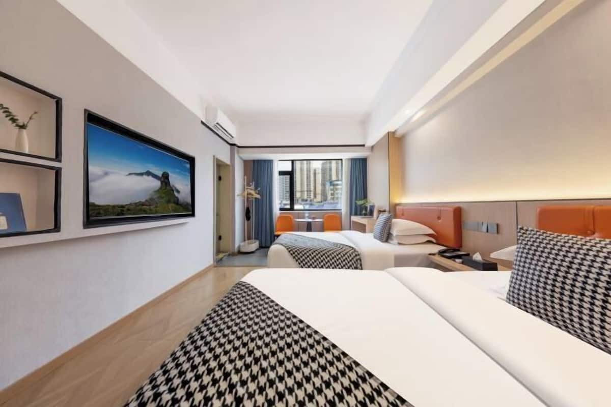Hotellihuone, jossa on kaksi sänkyä, suuri ikkuna, josta on näkymä kaupunkiin, seinällä oleva taulu ja pieni pöytä tuolilla.