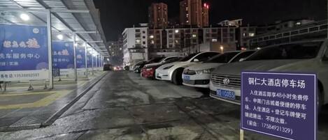 Un estacionamiento de noche con coches aparcados en filas, un cartel con texto y edificios al fondo.