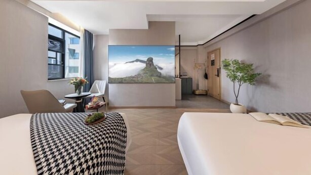 Una habitación de hotel con un ventanal grande, una cama, un escritorio con una silla, una planta en maceta y un cuadro en la pared.