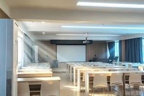 Uma sala de conferência com várias mesas e cadeiras, uma tela de projetor e um grande janelão com cortinas.