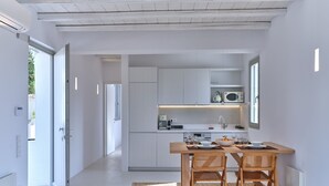 A modern kitchen with white cabinetry, a wooden dining table with two chairs, and a view of the outdoors through a glass door. - Perivoli Luxury Homes, Paros, Greece (Paros)