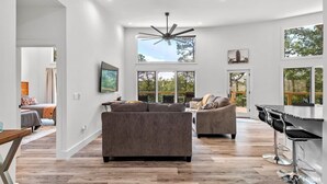 A spacious living room with a large ceiling fan, a dining area with a bar, and a view of the outdoors.