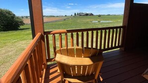 Una terraza de madera con una silla de madera, con vistas a un campo cubierto de césped y una casa a lo lejos.