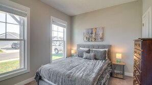 A bedroom with a large window, a bed with a gray and white striped bedspread, two bedside lamps, a cityscape painting on the wall, and a dresser with drawers.