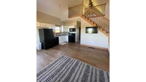A spacious living area with a wooden staircase, a kitchen with modern appliances, and a large rug on the floor.