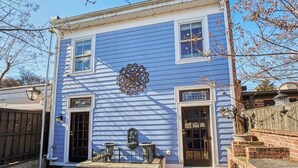 Una casa de dos pisos con fachada azul, un adorno metálico sobre la puerta principal y un pequeño porche con dos macetas de piedra.