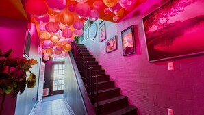 Una escalera con faroles colgantes rosa y naranja, un muro de ladrillo con cuadros enmarcados, y una puerta al final.