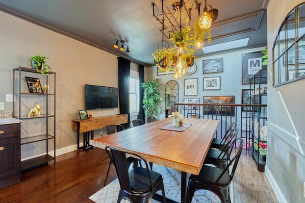 A dining area with a wooden table, black chairs, and a chandelier. There is a TV on the wall, a bookshelf, and a plant on the windowsill.