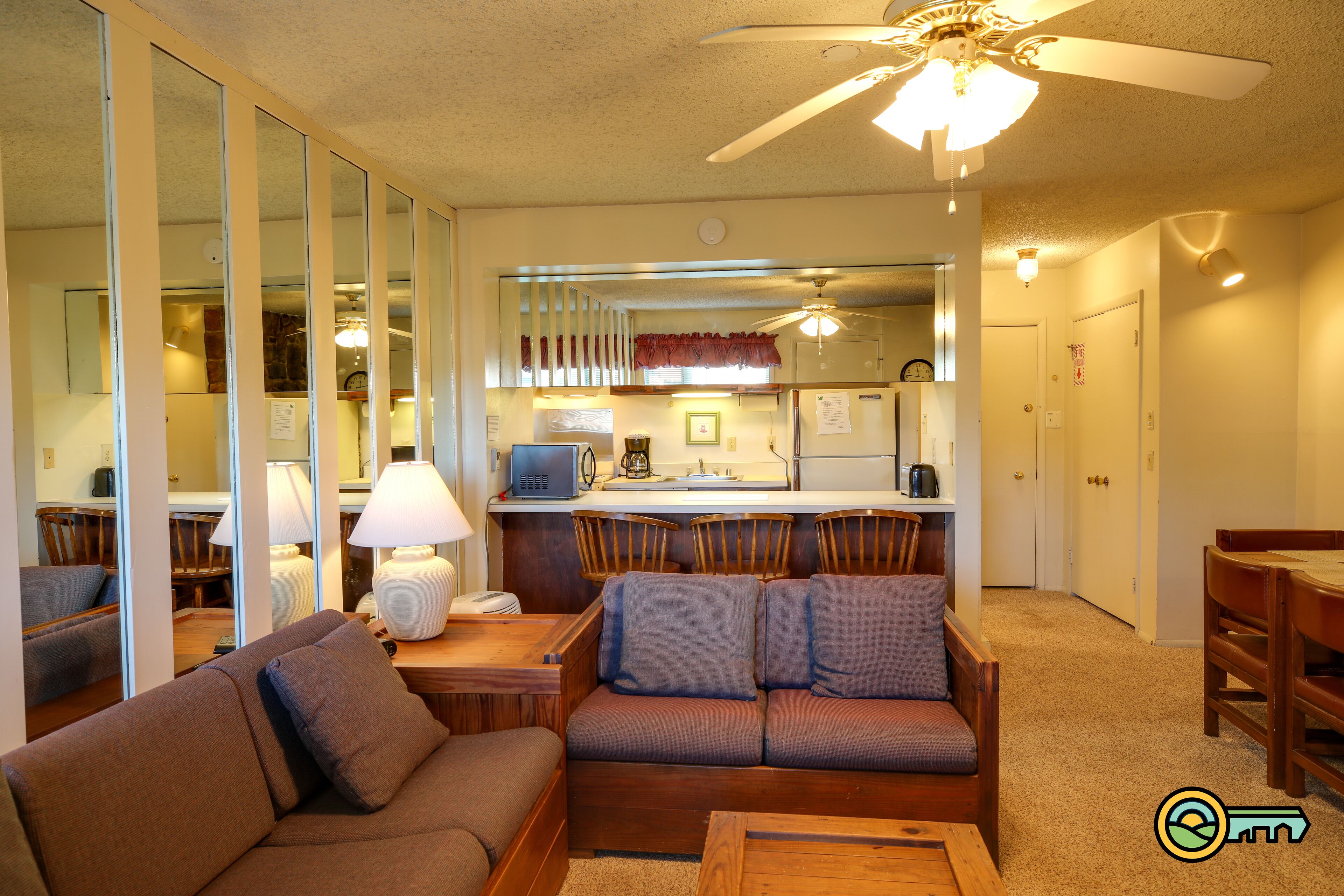 A living room with a sofa, a coffee table, and a dining area with chairs. There is a kitchen area with a refrigerator, stove, and microwave. A ceiling fan is visible above the kitchen.