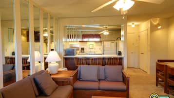 A living room with a sofa, a coffee table, and a dining area with chairs. There is a kitchen area with a refrigerator, stove, and microwave. A ceiling fan is visible above the kitchen.