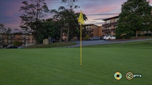 Een golfbaan met een vlaggenstok op de voorgrond, omringd door bomen en woongebouwen in de verte.