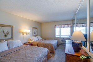 A hotel room with two beds, a desk with a lamp, a window with curtains, and a mirror.