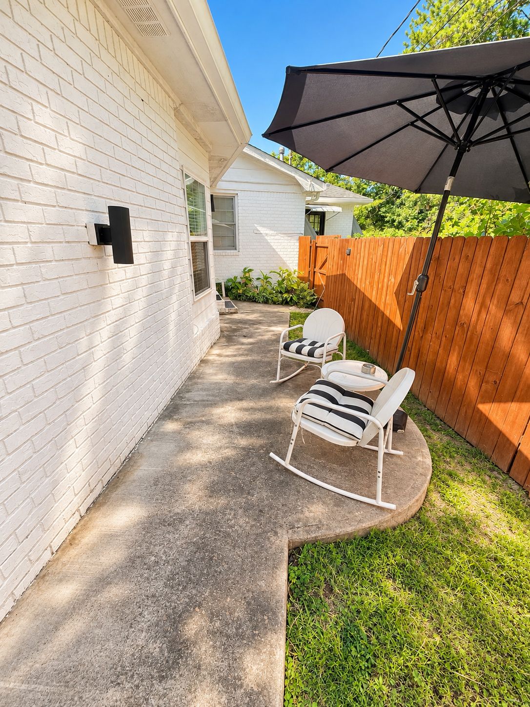 A patio area with a white brick wall, a wooden fence, a concrete pathway, and a rocking chair with a striped cushion.