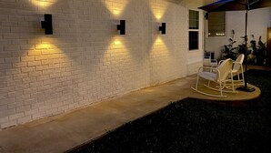 Een gevel van witte baksteen met drie wandlampen, een betonnen terras en een schommelstoel onder een blauwe parasol.