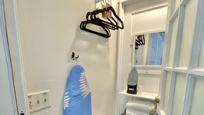 Een kleine, smalle kamer met een houten vloer, een blauw strijkplank, een zwarte stofzuiger en een deur met een bordje.