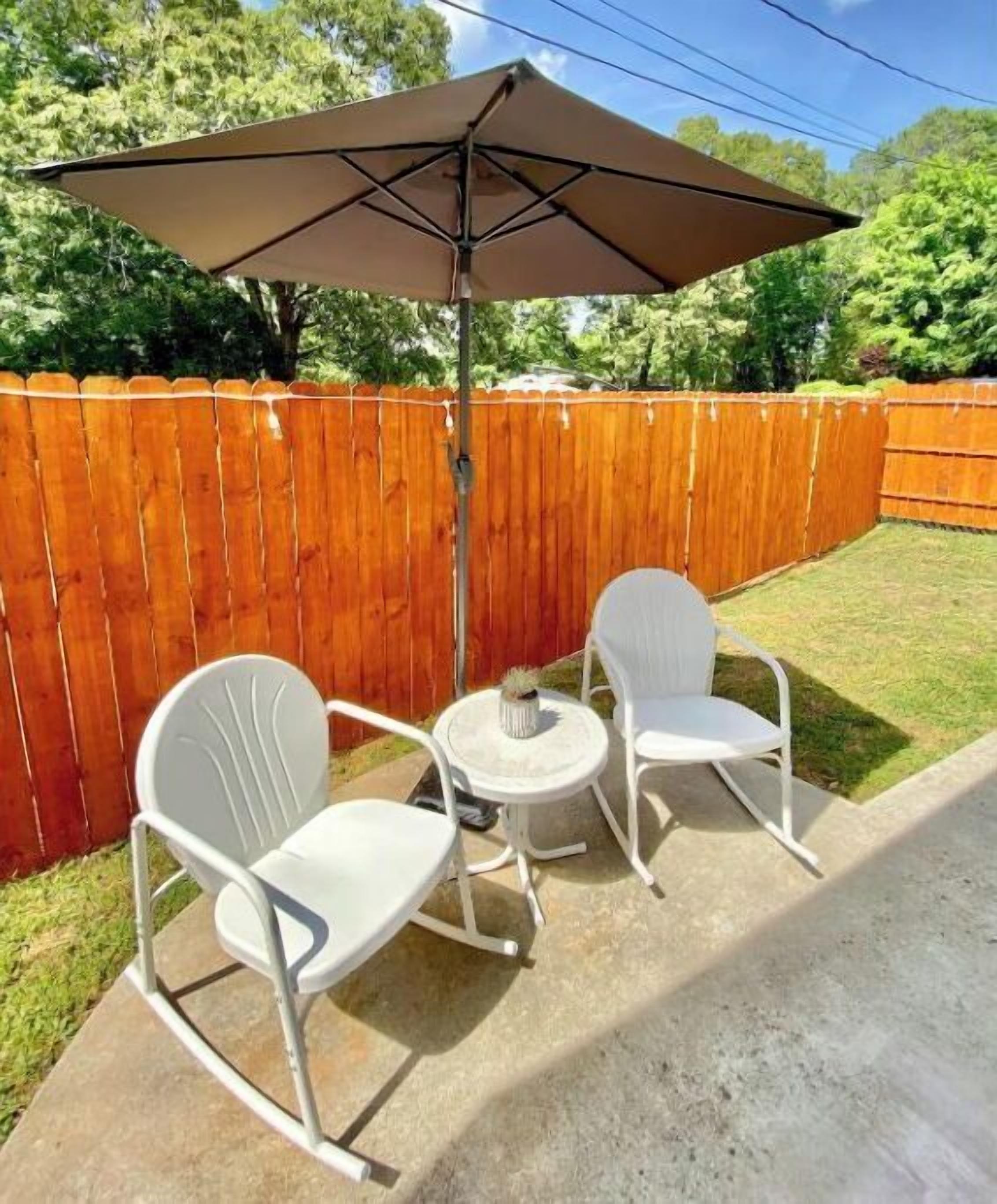 Un patio exterior con dos mecedoras blancas y una pequeña mesa redonda, cubierto por una gran sombrilla.