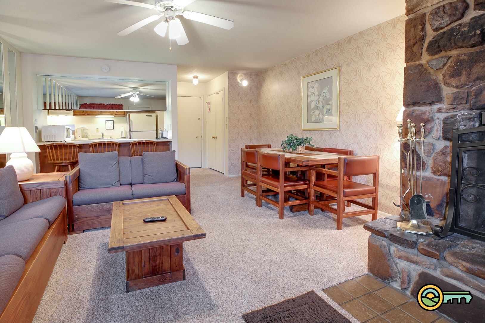 A cozy living room with a sofa, coffee table, and dining table set. A fireplace is visible on the right, and a kitchen area is in the background.