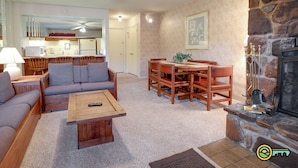 Ein gemütliches Wohnzimmer mit Sofa, Couchtisch und Esstisch. Rechts ist ein Kamin zu sehen, im Hintergrund befindet sich eine Küchenzeile.