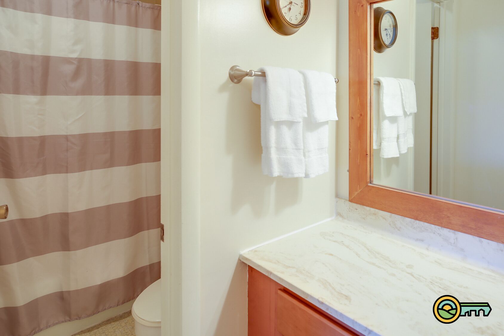 A bathroom with a striped shower curtain, a white toilet, a marble sink with a wooden cabinet, and two wall clocks.