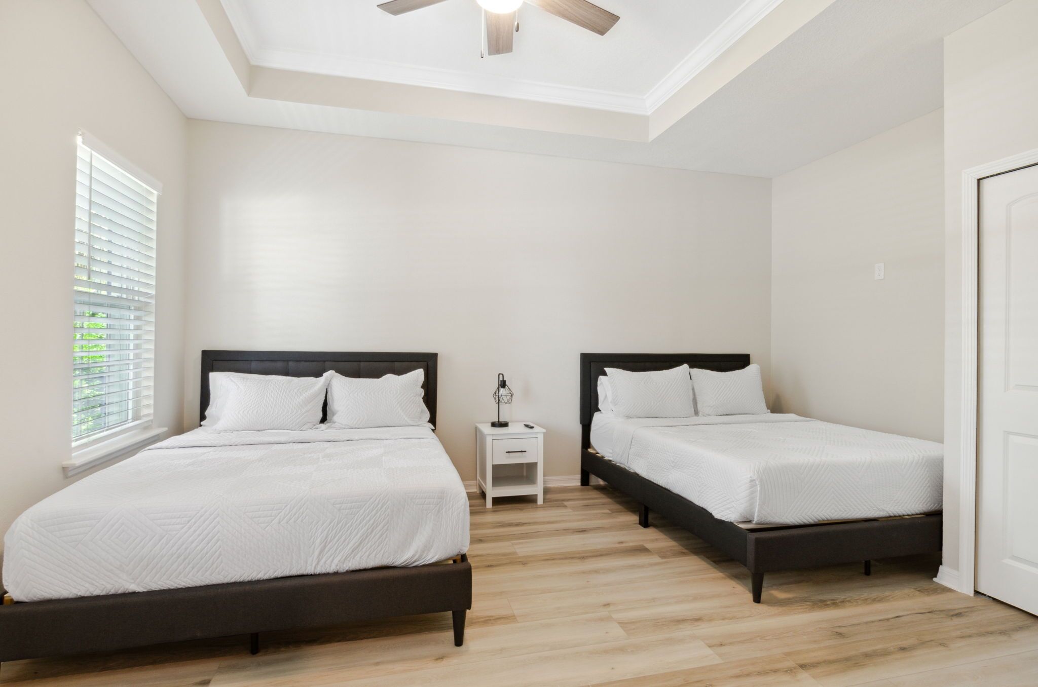 A bedroom with two beds, a ceiling fan, a nightstand, and a window with blinds.