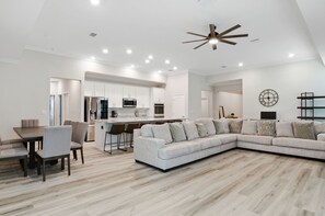 A modern living room with a sectional sofa, dining table, and chairs. There is a kitchen area with white cabinets and a microwave.