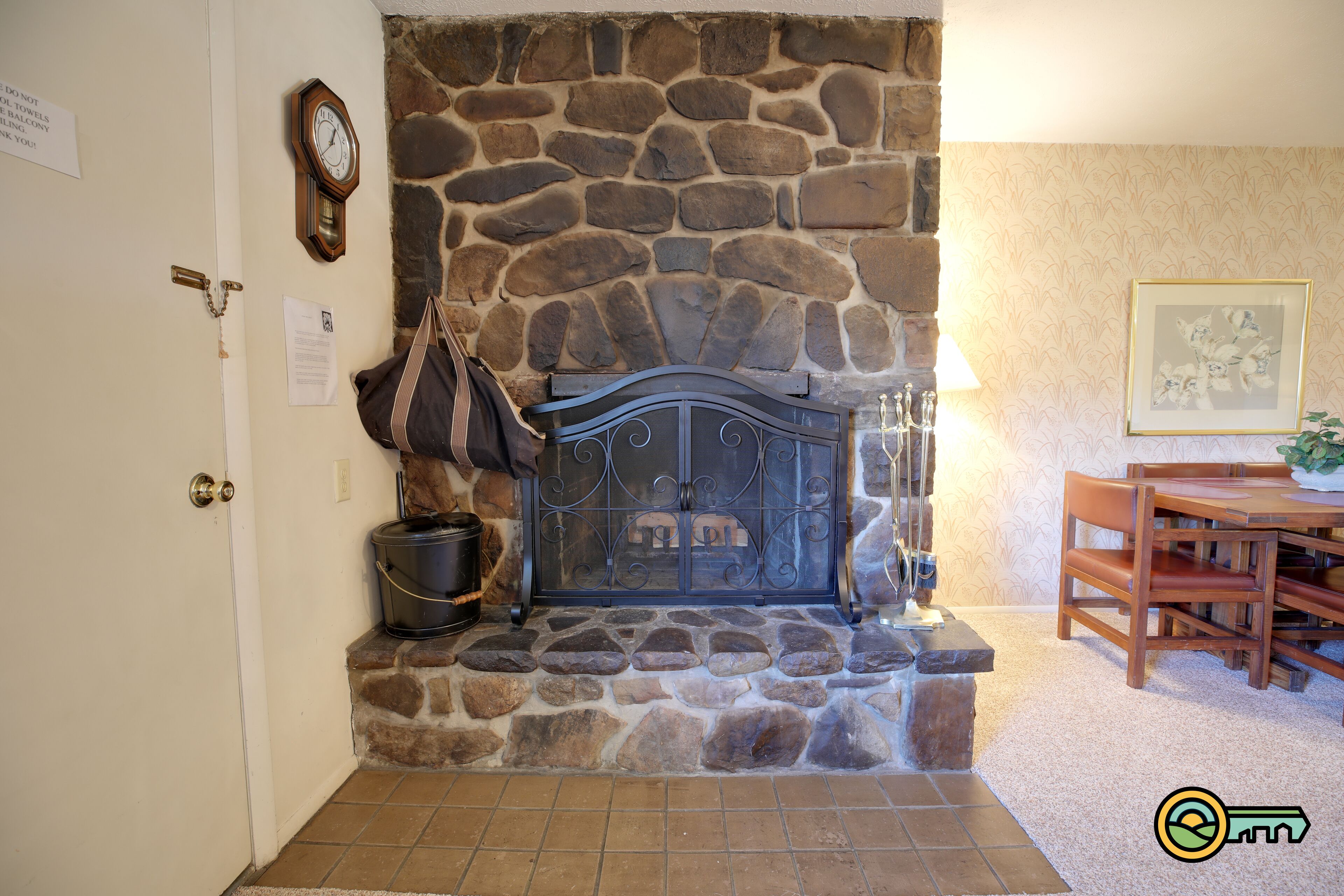 Una chimenea de piedra con rejilla de metal, un reloj en la pared y un comedor con sillas y mesa de madera.