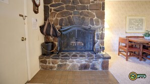 Lareira de pedra com grelha de metal, um relógio na parede e um espaço de jantar com cadeiras de madeira e uma mesa.