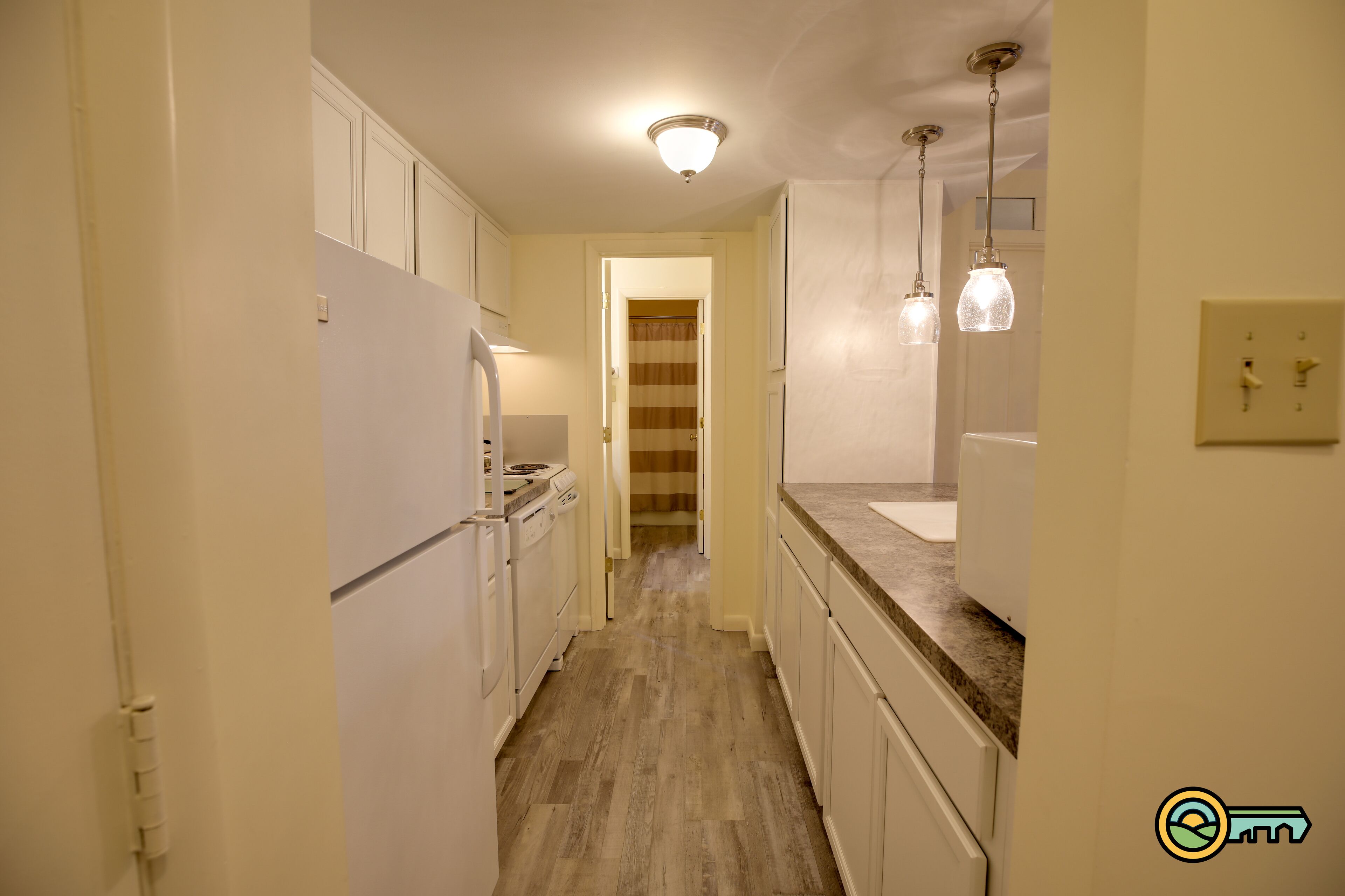 Una cocina estrecha con armarios blancos, suelo de madera y luces colgantes.