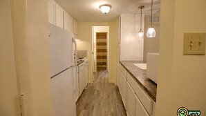 Une cuisine étroite avec des meubles blancs, un plancher en bois et des luminaires suspendus.