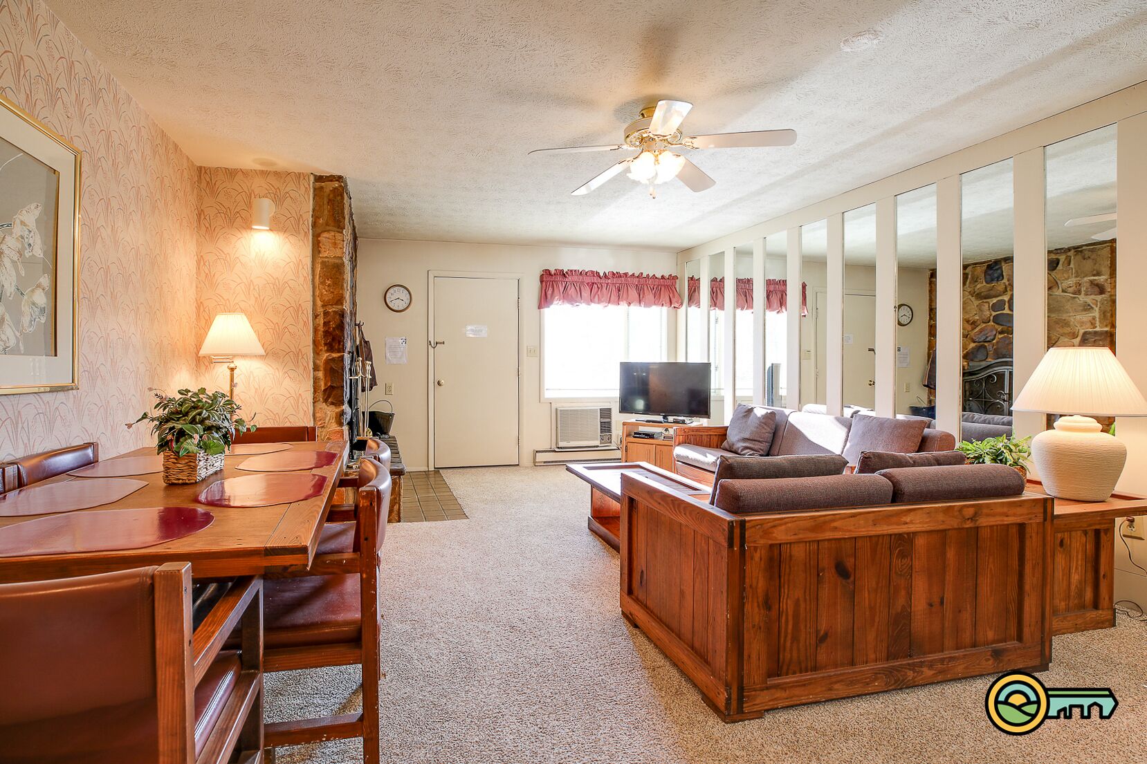 A cozy living room with a wooden coffee table, a sofa, and a television. There is a dining area with a table and chairs, and a stone wall in the background.
