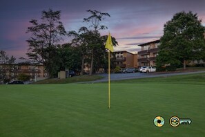 A golf course with a flagstick, surrounded by residential buildings and parked cars.