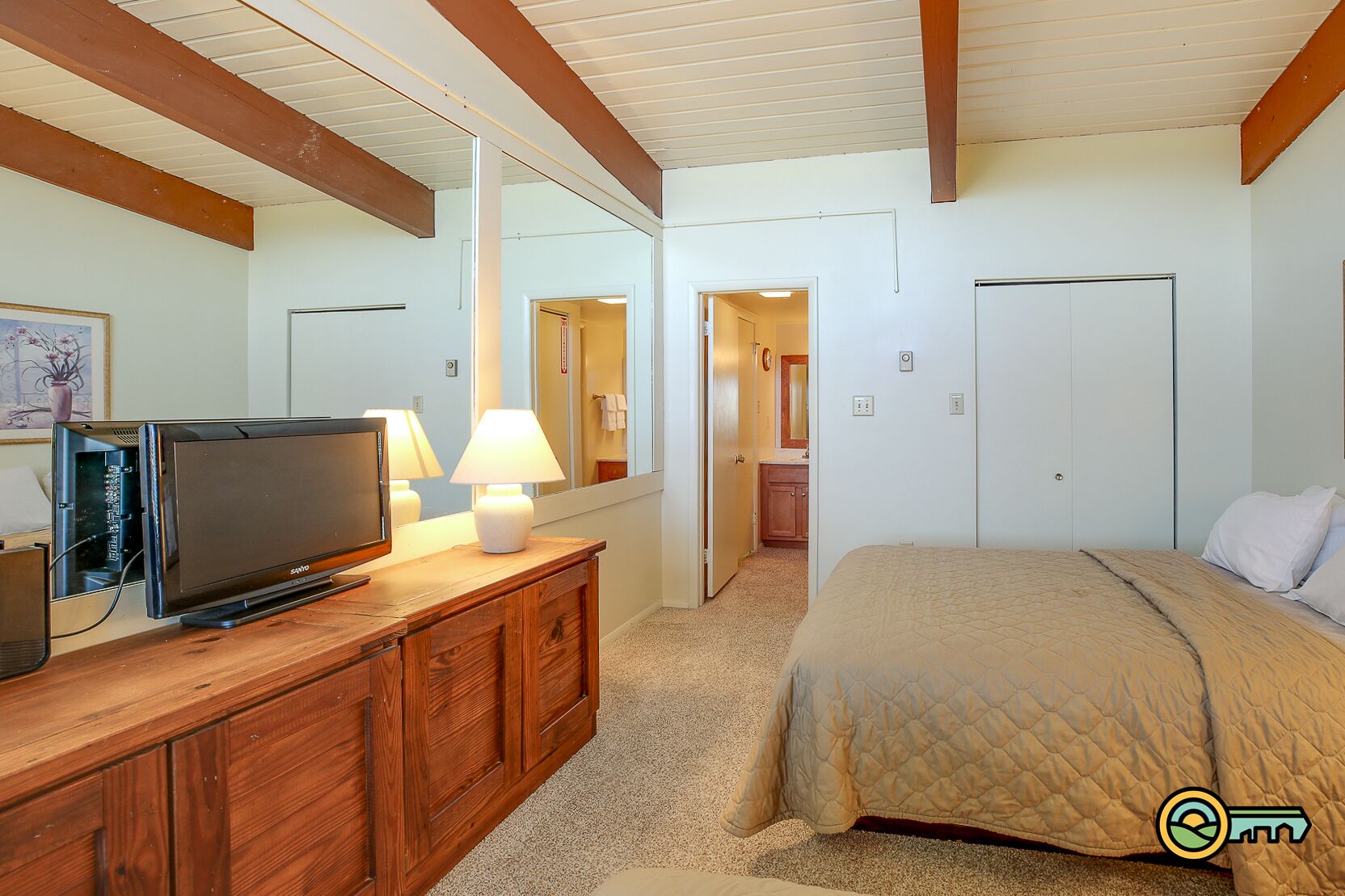 A bedroom with a bed, a TV on a wooden dresser, a mirror, and a door leading to a bathroom.