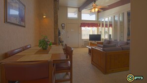 A dining area with a wooden table and chairs, a television, and a sofa in a living room with large windows and a ceiling fan. - Ski/MTB/Golf Condo (#132) - Slope side | Mountain, golf course views | Vaulted ceilings! (Basye)