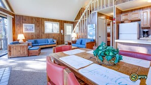 A cozy cabin interior with a dining area, blue sofas, wooden walls, and a kitchen.