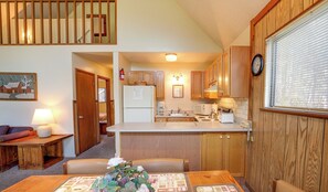 A compact kitchen with wooden cabinets, a white countertop, and basic appliances. There is a dining area with a table set for two, a lamp, and a flower centerpiece.