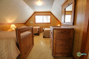 A cozy attic bedroom with wooden walls, a large window with blinds, a wooden dresser, and two beds.