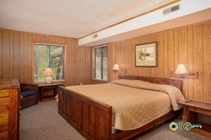 A bedroom with a wooden paneled wall, a bed with a beige quilt, two bedside lamps, a desk with a chair, and a framed picture on the wall.