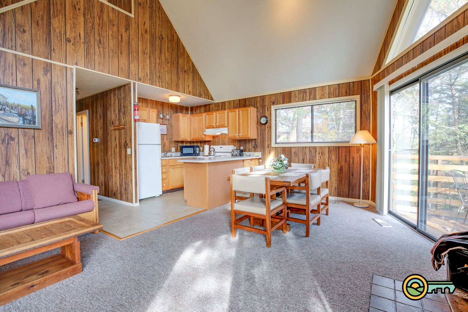 A cozy cabin interior with a wooden interior, a dining area with a table and chairs, a kitchen with a refrigerator and microwave, and a large window providing natural light.