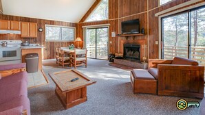 A spacious living room with a fireplace, wooden furniture, and large windows offering a view of the outdoors.