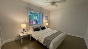 Un dormitorio con una cama, dos mesitas de noche con lámparas, un ventilador de techo, un ventanal con cortinas y un cuadro colgado en la pared.