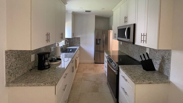 Una cocina moderna con armarios blancos, encimeras de granito, electrodomésticos de acero inoxidable y una isla central con fregadero y hervidor.