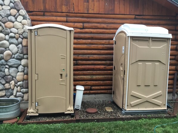 Two portable toilets in beige, one with a door and the other with a lid, placed outdoors against a wooden wall with a stone border.